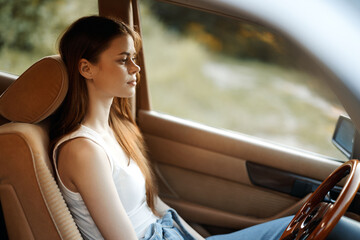 Fototapeta premium A young woman with long brown hair sitting in a car, expressing calmness and contemplation, surrounded by nature with soft sunlight filtering through, reflecting a serene lifestyle.