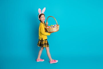 Happy young girl with bunny ears holding a colorful Easter egg filled basket, smiling playfully in a cheerful pose