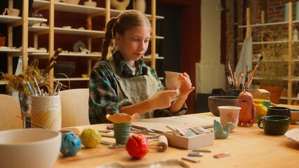 Adorable Caucasian little girl teenager child daughter school kid pottery sculptor artist potter examine inspecting clay craft vase handmade artwork crockery creative stoneware inside workshop studio