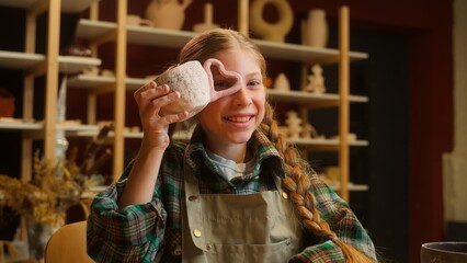 Smiling happy Caucasian little girl school child kid schoolgirl student potter artisan having fun during pottery class lesson looking at camera through heart shape cup handle indoors workshop ceramic
