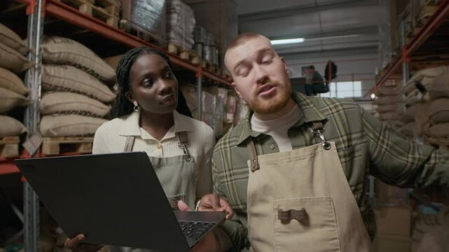 Man and African American woman in aprons holding laptop and collecting data for inventory audit in large storage area of coffee roasting plant