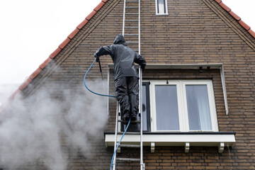 Man on ladder in waterproof suit cleans dirty brick facade of house with high pressure water jet....
