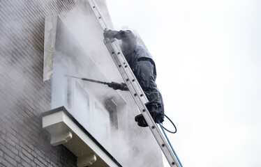 Man on ladder in waterproof suit cleans dirty brick facade of house with high pressure water jet....