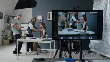 Studio monitor displaying live feed of actress at desk having her make-up adjusted, man walking by with papers, amidst professional lighting and cameras