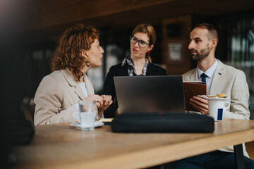 Three professionals sitting at an outdoor cafe, engaging in a business discussion and collaboration, with coffee cups and a laptop present.