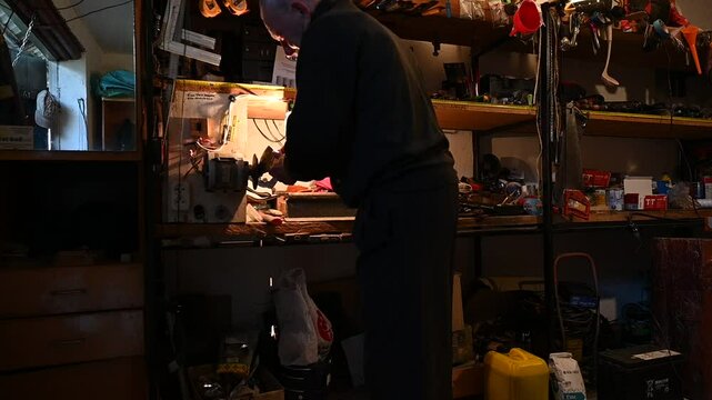 Grandpa sharpens an axe in his workshop. A dedicated craftsman is focused on repairing equipment in a small workshop filled with various tools and materials.