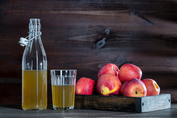 Apple cider in glass bottle and glass next to fresh red apples in wooden tray on kitchen table, closeup, copy space
