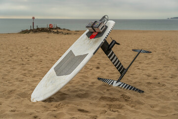 Electric powered hydrofoil surf board with deck mounted battery.  Sandy beach, sea and overcast sky...