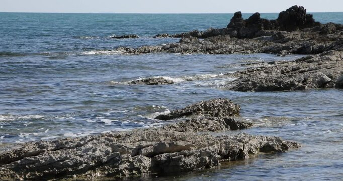 Wide shot of ocean waves splashing against a rocky shore, with a uniquely shaped rock formation resembling a camel toe in the distance. The dramatic seascape captures nature&rsquo;s raw power and beauty.
