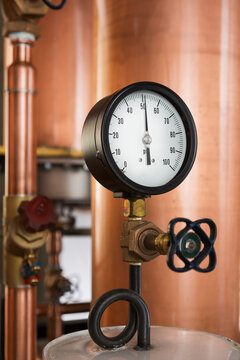 A copper still in a distillery, a large copper chamber and pipework and a large gauge and tap.