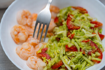 Freshly made salad with chopped napa cabbage, red bell pepper, avocado dressing and boiled shrimps is served on a white plate with a fork reaching for it