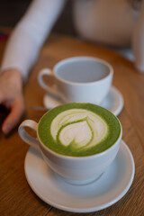Steamed milk creates a heart shape design in a matcha latte, resting on a wooden table alongside another cup, creating a cozy cafe atmosphere