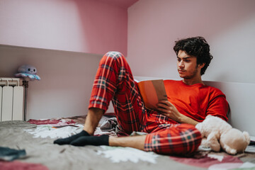 A young man in red pajamas focuses on reading a book while relaxing on his bed. The setting...