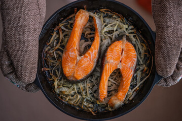 Chef wearing oven mitts is holding a cast iron skillet containing two roasted salmon steaks on a...