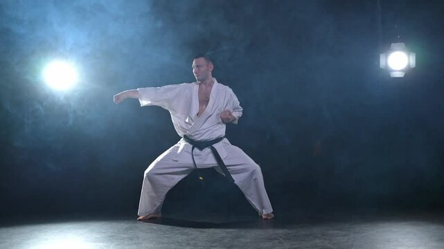 Strong man fighter in white kimono for karate martial arts with black belt preparing for fight training in black background. Man training moves punches kicks practicing shadow fight ready for sparring