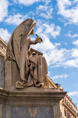Side of old marble statue located in historic square in Rome representing a bearded man covered by a hood holding a pen is viewed against a blue sky with clouds