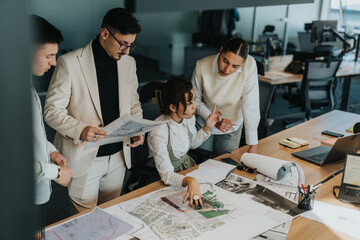 Obraz premium A diverse group of business colleagues gathered around a table, engaged in discussing and analyzing architectural documents and plans for a project in a modern office setting.