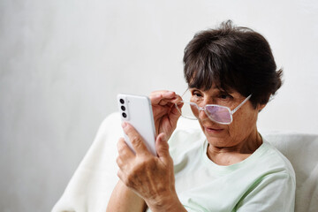 Confused elderly woman in eyeglasses has problem with bad vision, blurry sight reading on mobile...