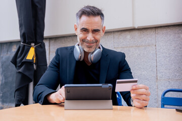 Confident caucasian man with headphones smiling while using tablet and holding a credit card...