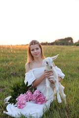 A young woman in a white dress sits in a field with a white baby goat on her lap and a bouquet of...