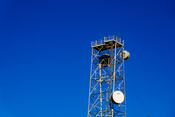 Infrastructure, a tall metal communication tower with a platform at the top, satelite and receiver dishes on the side, against a blue sky.