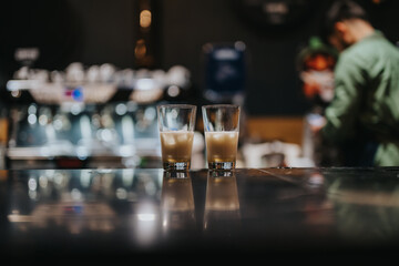 One glass stands prominently on a polished counter, with another beside it, amid soft lighting and bokeh effects from a professional coffee machine, featuring a barista in blurred motion in the