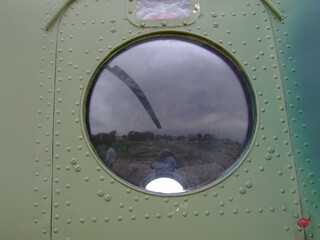 Round window on green military helicopter fuselage reflecting landscape and rotor blade