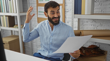 Hispanic man with beard laughing while reading documents in a bright office with shelves and files...
