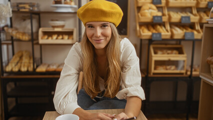 Young, blonde, woman wearing a yellow beret smiling in a cozy bakery with shelves full of fresh bread and pastries indoors