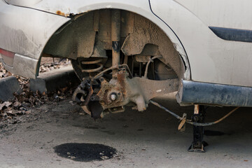 Old car with missing rear wheel stands on jack in urban yard, exposing rusty suspension and dirty...