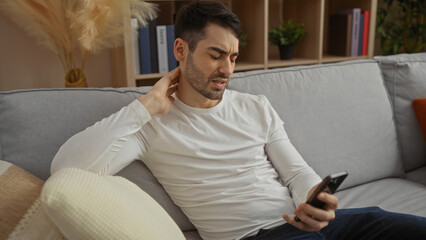 Young man sitting on a sofa in a living room, holding a smartphone with a pained expression, indicating discomfort, surrounded by a cozy home interior.