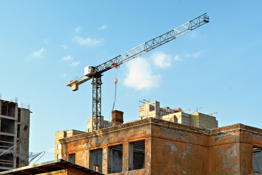 Tower crane operates above old crumbling brick building as new construction rises behind it under clear sky, symbolizing urban renewal and change. - Powered by Adobe