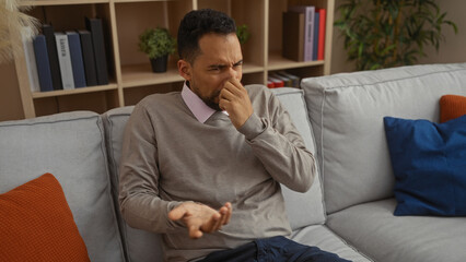 Young man in a living room covering nose with hand due to a bad smell, showcasing a reaction to an...