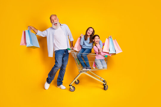 Happy grandfather with his two granddaughters enjoying shopping together in a trolley against yellow background
