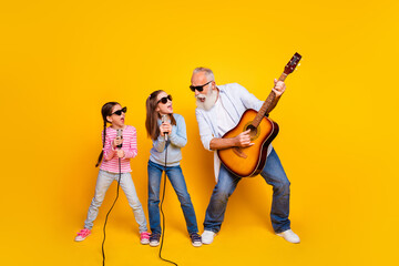 Cheerful grandfather playing guitar with two granddaughters singing on microphones against a lively...