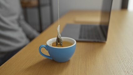 Blue mug with tea bag near laptop on wooden desk in modern office signifies relaxing work environment and productivity.