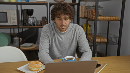 Young man working in office wearing casual sweater with coffee and donut on desk, looking focused while using laptop in organized workspace with shelves and kitchen items visible