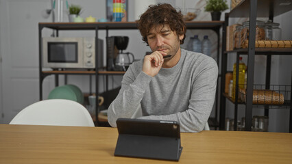 Fototapeta premium Young man thinking in office setting while sitting at a desk with tablet and holding pen, surrounded by shelves with kitchen items.