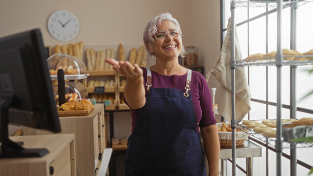 Smiling elderly woman in a bakery extending her hand to customers surrounded by bread and pastries wearing a purple shirt and apron indoors under a wall clock