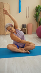 Obraz premium Hispanic elderly woman with grey hair doing yoga stretches on a mat indoors at a gym with various fitness equipment in the background showcasing a healthy, active lifestyle.