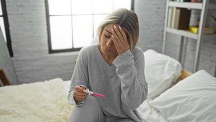 Woman sitting in bedroom holding pregnancy test looking worried with hand on forehead expressing concern in cozy home environment