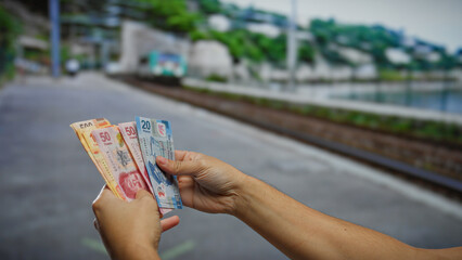 Man holding mexican pesos at an outdoor train station, symbolizing travel, finance, and investment against a blurred backdrop of tracks and nature.