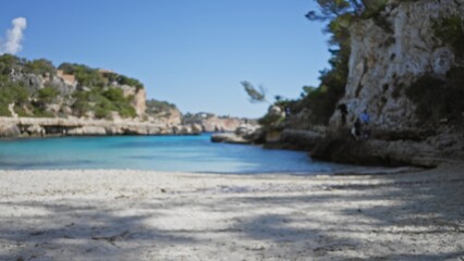 Blurred view of cala llombards beach in mallorca with defocused sand and clear water surrounded by rocky cliffs under a bright blue sky.