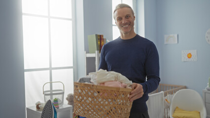Mature blonde caucasian man holding laundry basket indoors in a home laundry room setting, expressing friendly and positive demeanor.