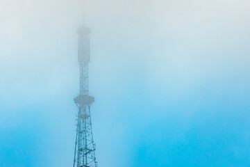 A tall communication tower emerges from thick fog against a faint blue sky, partially obscured by mist, creating a mysterious and atmospheric scene. Includes copy space.