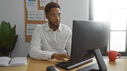 Young man sitting in an office working on a computer, in front of a window, with a notepad, and a red coffee mug on the desk