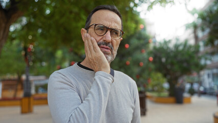 Middle-aged hispanic man in an urban park holding his cheek in pain, indicating a toothache amidst...