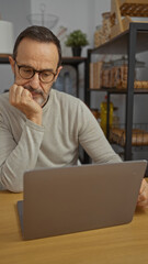 Mature man thoughtfully working on laptop at office desk in a modern workplace setting with shelves and decor visible