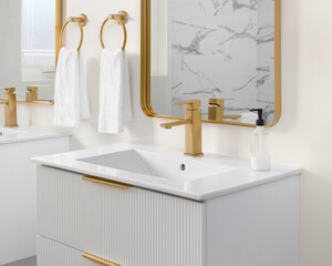 A bathroom detail with a white fluted floating cabinet, gold faucet on a white sink, and a gold mirror with a reflection of a marble tiled shower. © Joseph Hendrickson