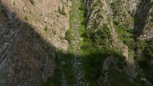 Aerial footage capturing a narrow, rocky chute on a mountain slope in Kyrgyzstan. The camera moves forward, revealing a steep, green valley with rugged cliffs and scattered vegetation.